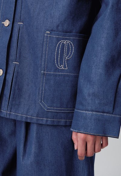 Primer plano de un bolsillo de chaqueta de mezclilla azul con un logo bordado en blanco y pantalones de mezclilla a juego, mostrando una mano relajada al costado.