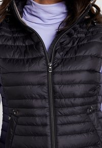 Czarna, pikowana kamizelka z wysokim kołnierzem, zamkiem błyskawicznym, bocznymi kieszeniami oraz charakterystycznymi poziomymi przeszyciami. Miękka, gładka tekstura.