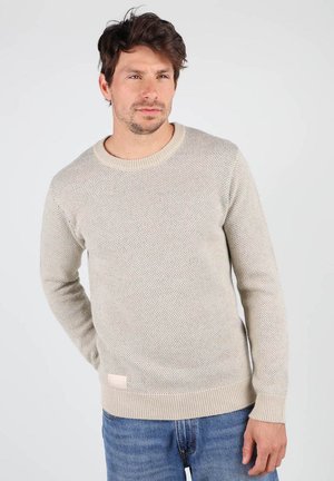 Pull en maille beige clair avec un motif texturé, col rond, poignets et ourlet côtelés. Associé à un jean bleu. Présente une petite étiquette sur le côté.