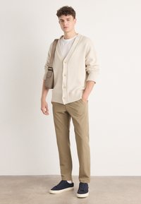 Cardigan bej tricotat cu decolteu în V, butoni mari și tiv côtat, asortat cu pantaloni bej și adidași bleumarin, ținând o geantă de umăr bej.