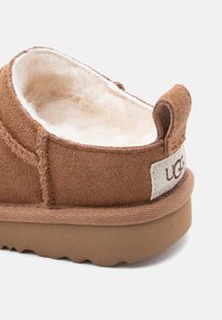 Καφέ σουέντ παντόφλα με μαλακή εσωτερική επένδυση από λευκή γούνα και παχύ υφή πέλμα, με υφασμάτινη θηλιά τραβήγματος και μερική ετικέτα μάρκας.