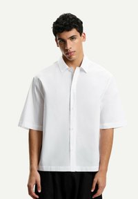 Camicia bianca a maniche corte con colletto a punta, chiusura con bottoni e una vestibilità rilassata. Texture liscia con un design minimalista.