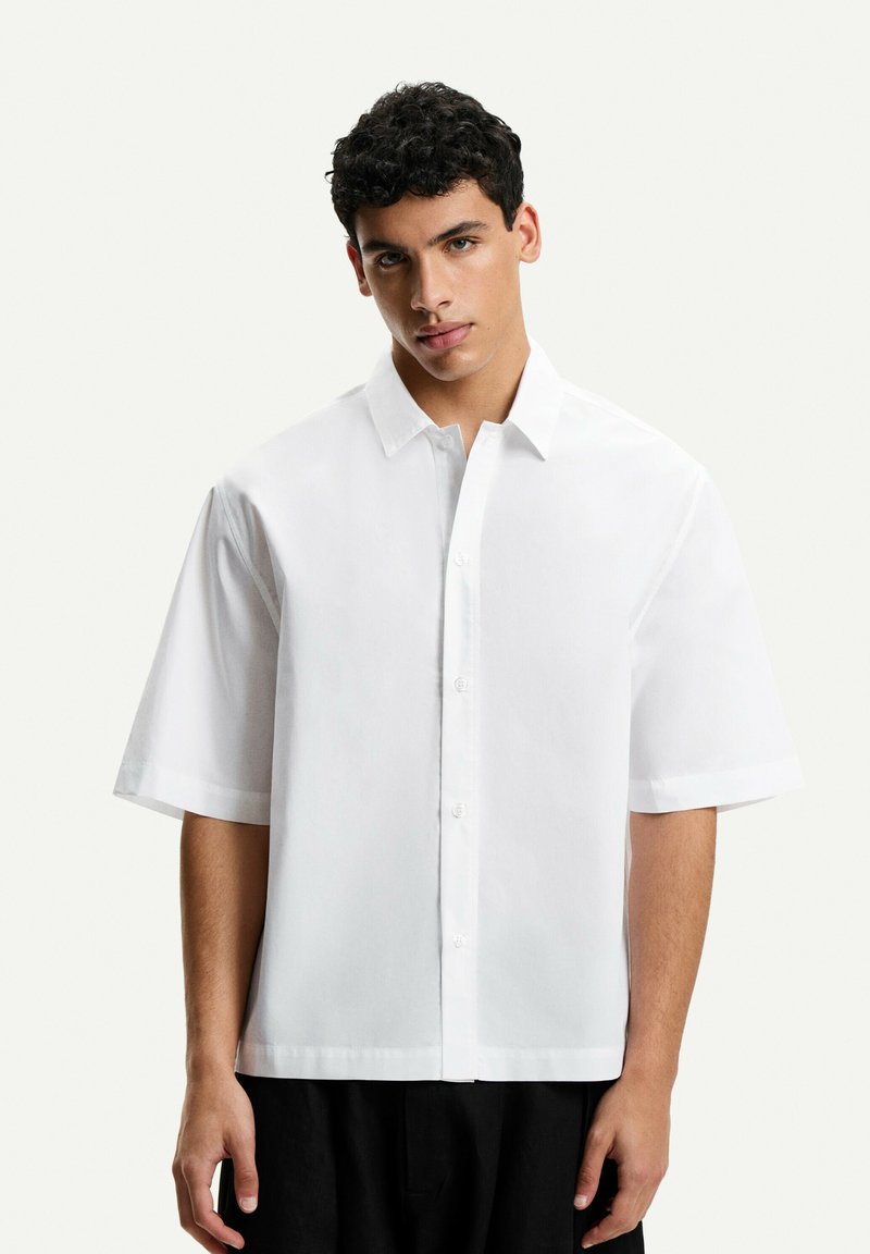 Camicia bianca a maniche corte con colletto a punta, chiusura con bottoni e una vestibilità rilassata. Texture liscia con un design minimalista.