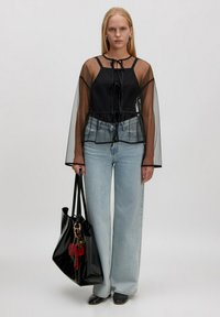 Frau mit glattem, blondem Haar, die ein-transparentes, schwarzes Langarmoberteil über einem schwarzen Tanktop, hellblauen weiten Jeans, schwarzen Stiefeln trägt und eine große schwarze Tote-Bag hält.