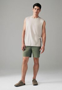 Mouwloos beige shirt, groene shorts en bruine instap schoenen op een staande mannelijke model tegen een effen achtergrond. Vlakke, soepele texturen zichtbaar.