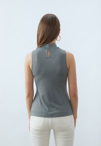 Grå tanktopp med høy krage og bak nøkkelhulldetalj, laget av glatt stoff. Kombinert med hvite, tettsittende bukser.