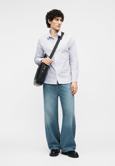 Νεαρός με κατσαρά μαλλιά που φοράει γαλάζιο πουκάμισο, τζιν παντελόνι με φαρδιά γραμμή, μαύρα παπούτσια και κρατά μαύρη τσάντα χιαστί, κοιτάζοντας προς το πλάι.