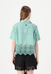Muntgroene kantblouse met een gedetailleerde geschulpte zoom, korte mouwen en een accent op de achterkant van de kraag, gecombineerd met zwarte shorts met kantafwerking.