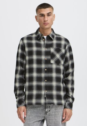 Ung mand iført en sort- og hvidternet skjorte med knapper og en forlomme, kombineret med lysegrå jeans, stående mod en ensfarvet baggrund.