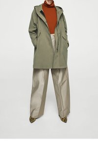 Vêtements d'extérieur vert olive avec capuche, présentant des poches avant. Associé à un col roulé couleur rouille et à un pantalon beige large. Chaussures texturées.