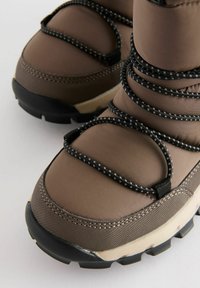 Bruine geïsoleerde laarzen hebben een textuuroverlay, zwarte elastische veters en een robuuste zool voor grip. Het ontwerp legt de nadruk op warmte en stabiliteit.