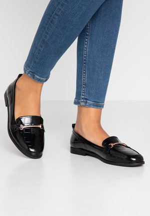 Mocasines de charol negro con textura de cocodrilo, suela plana y acentos en dorado. Llevados con jeans azul oscuro, mostrando el tobillo.