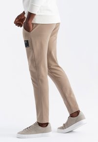 Beige avsmalnande byxor i mjukt tyg, med en sidficka och en svart etikett. Matchas med ljusgrå sneakers för en avslappnad stil.