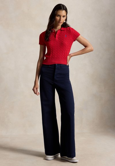 Žena stojaca s rukou na boku, oblečená v červenom krátkym rukávom polopulóvri s pleteným vzorom, tmavomodrých širokých nohaviciach a bielych teniskách na béžovom pozadí.