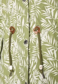 Grön jacka med ett vitt bladmönster, trädetaljer och flätade dragsnören. Innehåller tryckknappar och har en mjuk textur.