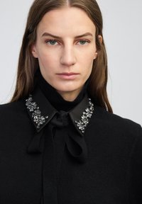 Vrouw met lang bruin haar, gekleed in een zwarte trui met hoge kraag en een zwart shirt met een juweelachtige kraag, vastgebonden met een strik.