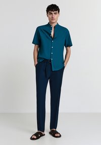 Türkises kurzärmliges Hemd mit einem knöpfbaren Kragen, kombiniert mit navyfarbener Hose und schwarzen Sandalen. Das Outfit hat eine moderne, maßgeschneiderte Passform.