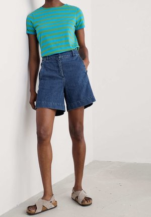 Osoba ubrana w niebiesko-zieloną, pasiastą koszulkę, ciemne szorty dżinsowe z wysokim stanem oraz beżowe sandały z odkrytymi palcami, stojąca przy białej ścianie.