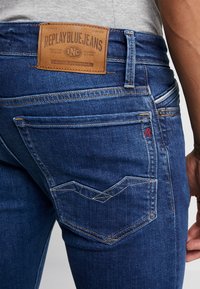 Dżinsy w ciemnoniebieskim kolorze z jasnym szyciem, z skórzaną naszką oznaczoną "Replay Blue Jeans" z tyłu oraz charakterystycznym wzorem kieszeni.