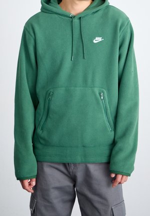 Person iført en grøn Nike fleece-hættetrøje med lynlåslommer foran og grå cargobukser, stående mod en ensfarvet lyse baggrund.