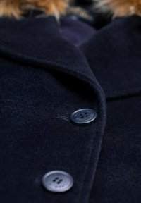 Cappotto di lana navy con una trama morbida, caratterizzato da un colletto su misura, due grandi bottoni e un bordo in pelliccia sintetica sul collo.
