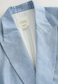 Světle modrý lněný blazer s klopou, strukturovanými klopami a pruhovanou podšívkou. Na štítcích jsou uvedeny informace o materiálu a značce.