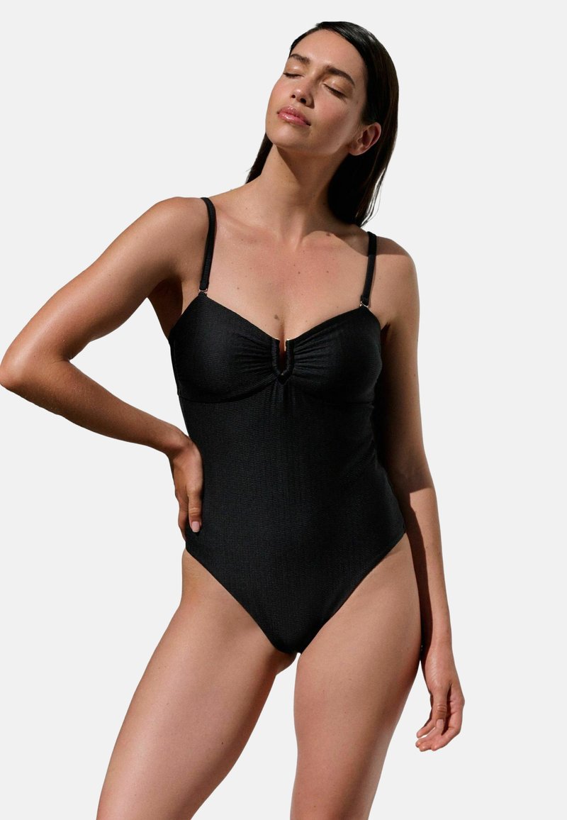 Femeie purtând un costum de baie întreg negru cu bretele subțiri, stând cu o mână pe șold și cu ochii închiși, pe un fundal simplu.