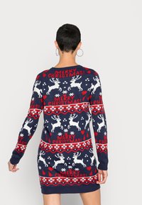Personne aux cheveux courts portant une robe-pull de Noël bleu marine avec des rennes blancs et le texte rouge "Joyeux Noël", vue de dos.