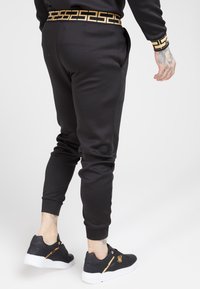 Svarta joggers med slim fit, som har ett midjeband och ärmslut med guldmönster. Matchas med svarta sneakers med guldakcenter.