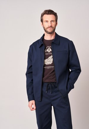 Man met baard, gekleed in een marineblauw jack met rits en bijpassende broek over een donker grafisch T-shirt, staand tegen een effen lichte achtergrond.