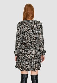 Femme aux longs cheveux roux portant une robe à manches longues imprimée léopard et des bottes noires montant jusqu'aux genoux, vue de dos sur un fond uni.