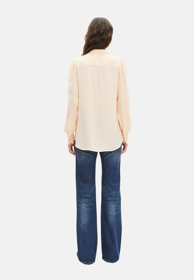 Mujer con cabello largo y castaño, vistiendo una blusa de manga larga color beige claro y jeans azul oscuro, de pie mirando hacia atrás sobre un fondo liso.