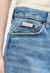 Κοντινό πλάνο τσέπης από τζιν ανοιχτού μπλε με χαλκινούς κόμπους και λευκή ετικέτα που γράφει "Calvin Klein".