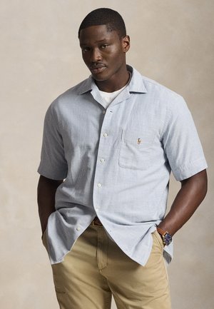 Άνδρας που φοράει ανοιχτό μπλε πουκάμισο με κοντά μανίκια πάνω από λευκό μπλουζάκι, μπεζ παντελόνι και σκούρο ρολόι χειρός, στέκεται μπροστά σε απλό φόντο.