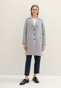 Manteau en laine gris avec un col à revers, trois boutons et des poches latérales, associé à un pantalon cropped bleu marine et des chaussures noires à lacets.