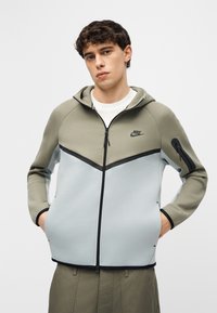 Jaunas vyras dėvi dviejų atspalvių pilką ir alyvuogių spalvos Nike užsegamą džemperį su kišenėmis rankose, stovintis priešais vienodą šviesų foną.