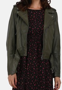 Woman wearing an olive green leather jacket over a black dress with small red floral print, shown from neck to hips.