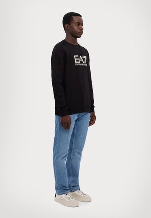 Felpa nera in cotone con logo, abbinata a jeans in denim azzurro chiaro e sneakers beige. Il modello è posizionato davanti a uno sfondo neutro.