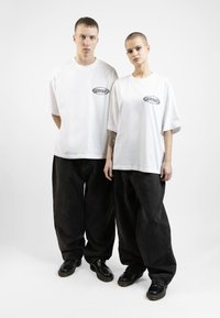 Two individuals wear oversized white cotton t-shirts with black logos, paired with loose-fitting black denim pants and black shoes.