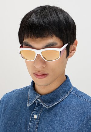 Giovane uomo con capelli corti neri che indossa occhiali da sole con montatura bianca e lenti arancioni e una camicia di jeans blu con bottoni.