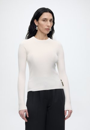 Γυναίκα με μακριά σκούρα μαλλιά που φορά ασημένια σκουλαρίκια, ένα εφαρμοστό λευκό μακρυμάνικο τοπ και άνετο μαύρο παντελόνι σε απλό λευκό φόντο.