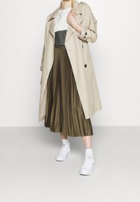 Trench coat beige con grandi bottoni, indossato sopra una maglietta grigia e bianca e una gonna plissettata verde oliva. Completato con sneakers bianche.