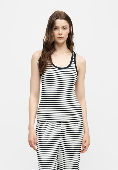Femme aux longs cheveux bruns portant un haut blanc sans manches et un pantalon à fines rayures horizontales noires, debout devant un fond uni.