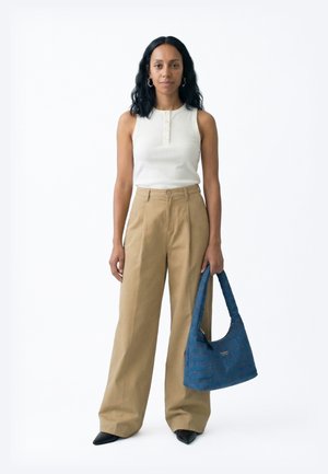 Hosszú fekete hajú nő fehér ujjatlan felsőben és széles bézs nadrágban, jobb kezében kék, texturált kézitáskával.