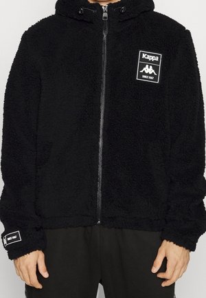 Czarna bluza polarowa zapinana na zamek z logo Kappa i tekstem "Since 1967" na piersi i na mankiecie lewego rękawa, noszona przez osobę od szyi do bioder.