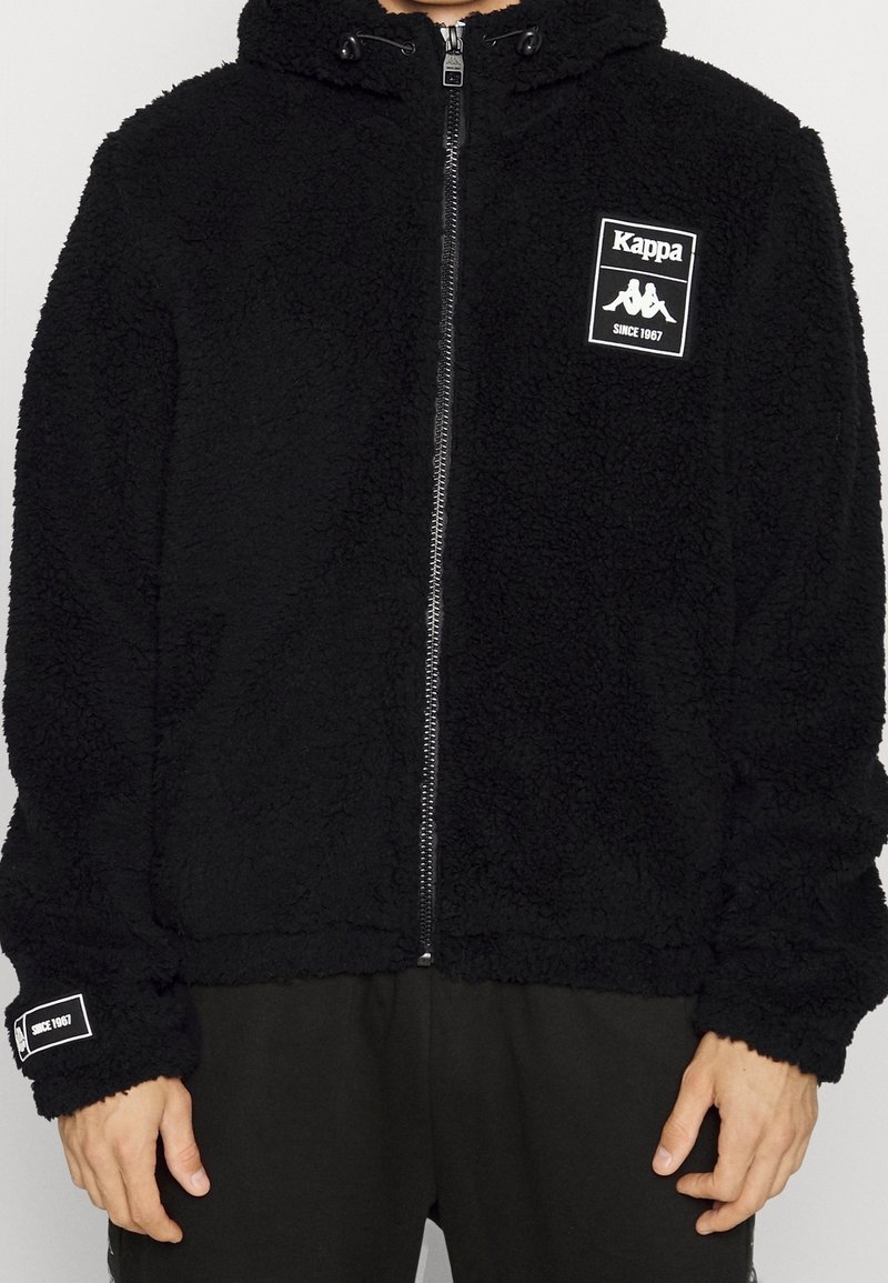 Veste polaire noire zippée avec le logo Kappa et le texte "Since 1967" sur la poitrine et le poignet de la manche gauche, portée par une personne du cou aux hanches.