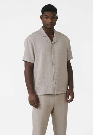Jasnobeżowa koszula na krótki rękaw na guziki, wykonana z miękkiego, teksturowanego materiału. Posiada kołnierz typu camp i luźny krój, zestawiona z pasującymi spodniami.