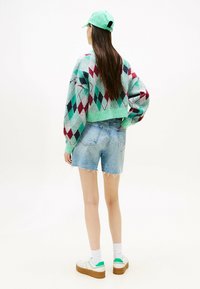 Maglione con un motivo a rombi verde e rosso, abbinato a pantaloni corti in denim azzurro e sneakers bianche con dettagli verdi.
