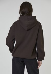 Persoon met lang donker haar, gekleed in een losse donkerbruine hoodie en zwarte broeken, met de rug naar de camera gekeerd tegen een effen lichte achtergrond.