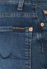Dunkelblaue Denim-Jeans mit einer Gesäßtasche und einem gestickten Logoetikett sowie kupferfarbener Hardware, die ein klassisches Design und eine ansprechende Textur zeigen.